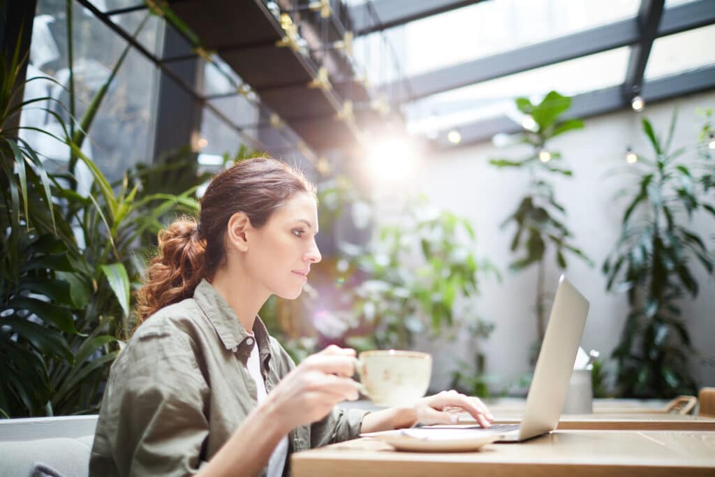 Serious concentrated young freelancer in casual shirt sitting at table and drinking coffee while using laptop and working on online project